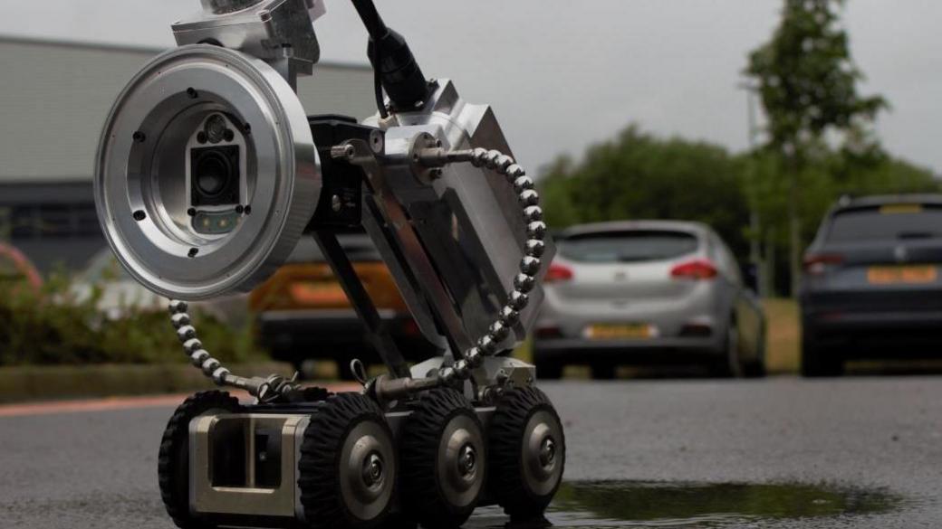 The silver robot has black wheels and is the fraction of the size of cars it is near in a car park.