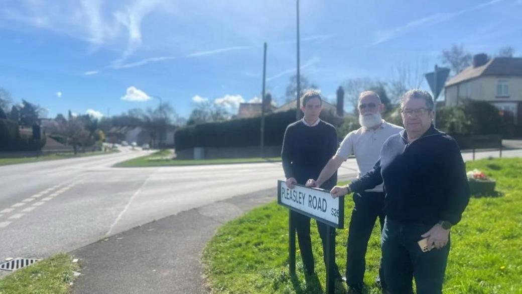 Three men stood behind a white road sign reading Pleasley Road with a road junction to the right of the image