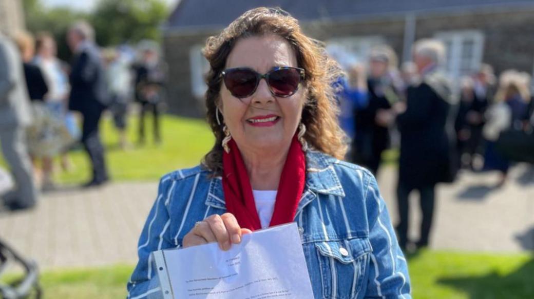 Millie Blenkinsop-French wears a blue and white denim jacket, red scarf and sunglasses. She has curly brown hair. She holds up a white document in a plastic wallet.