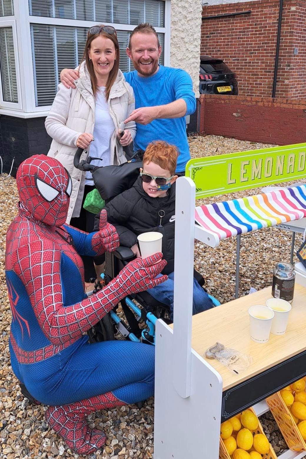 Thomas at his stand, as Spider-Man crouches next to him with a thumbs up and holding a cup of lemonade. Thomas's parents smile in the background, with his dad pointing at the superhero.