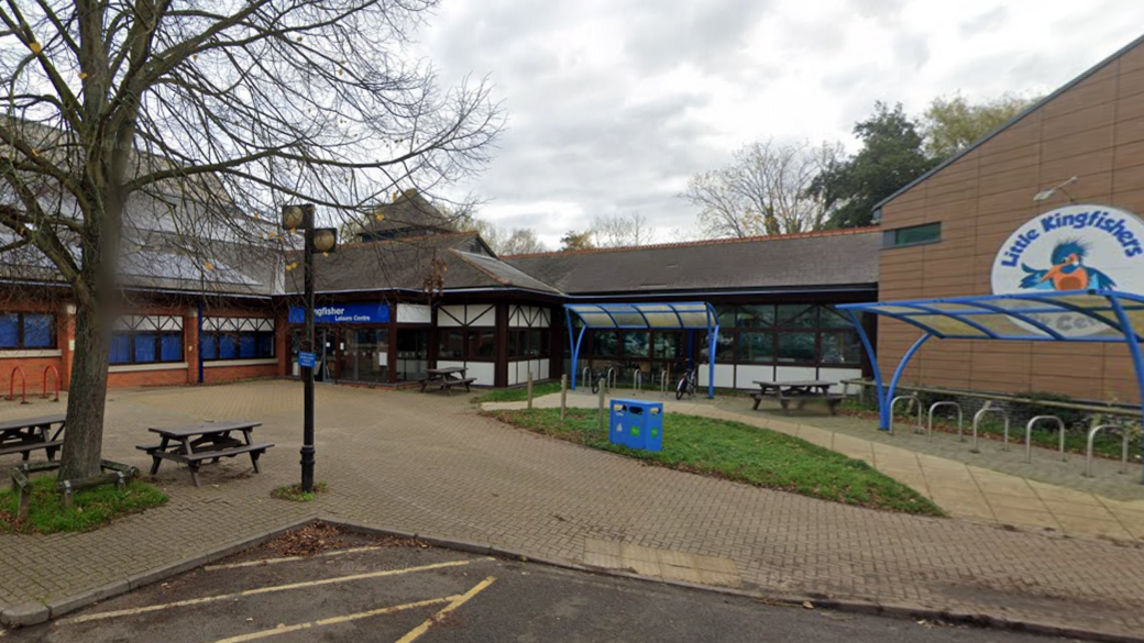 A bricked pool building to the left with a reception area in the centre of the photo. A large circular sign saying Little Kingfishers is on the right, with an empty bike rack in front of it. There is also a litter bin and two picnic benches in the foreground.