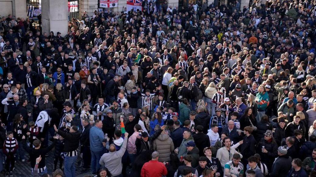 Overhead view of a gathering of Newcastle United supporters. The crowd consisting of many hundreds of people, some wearing Newcastle United football strips. Many have their heads thrown back and seem to be celebrating or chanting. 