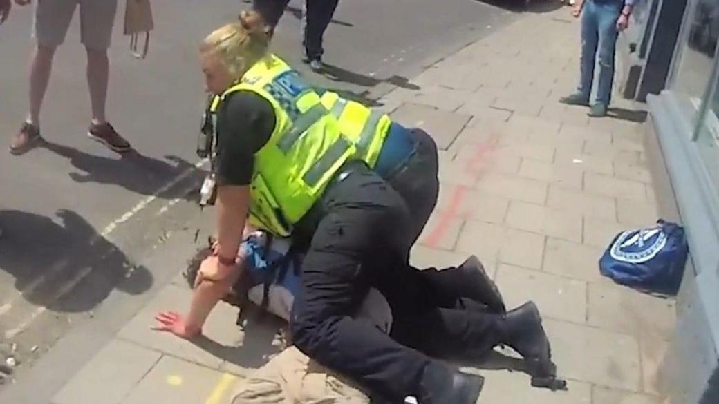 A still from a video of two police officers, both in uniform with high-vis vests are holding down a man on the pavement who has his arm out. People's legs are visible, indicating the public was aware and looking on.