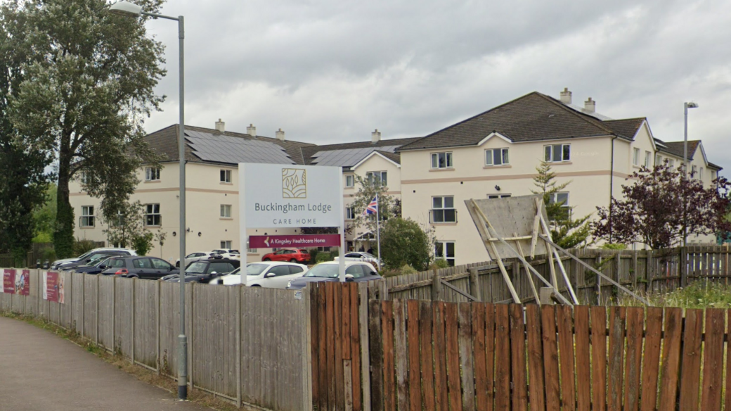 A view of Buckingham Lodge Care Home, in Watton, from the road. It is a cream-coloured building sitting behind a car park which is fenced off.