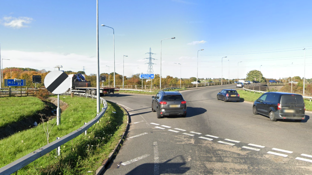 Cars are shown using a roundabout, with a grass verge to the left showing a national speed limit sign and a sign for the motorway ahead, with trees and an electricity pylon seen in the background.