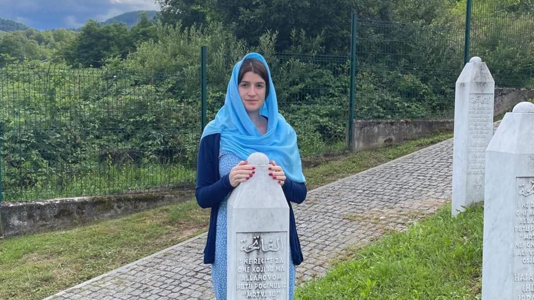 Una is standing next to her father's grave stone. She is wearing a blue dress and a blue hijab.