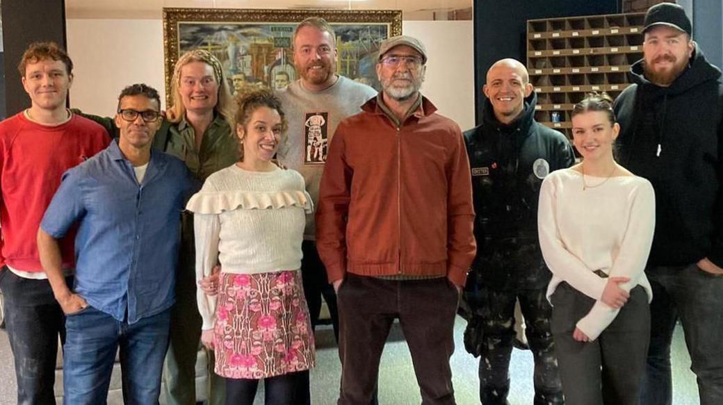 Artists gather around Eric Cantona in a studio he is wearing a flat cap and rusty brown jacket