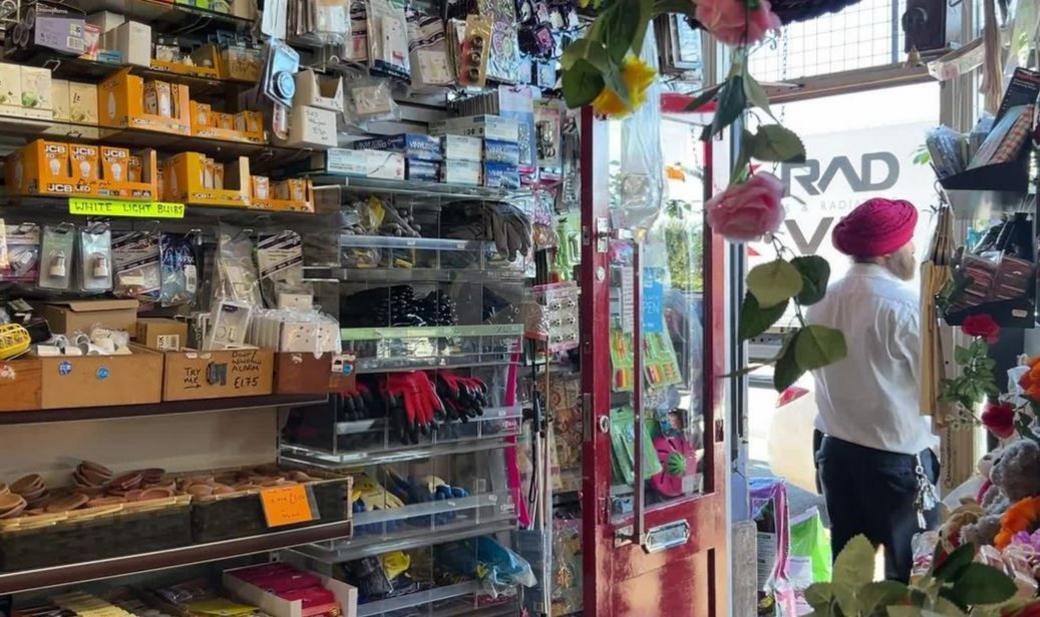 Interior of All Seasons DIY, with shelves of items like lightbulbs, screws and nails in the foreground, with Surinder Josan standing in the doorway looking into the street