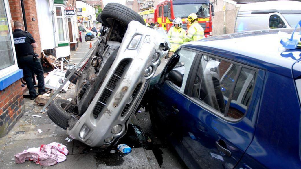 A silver car lies on its side on a pavement propped against a parked car