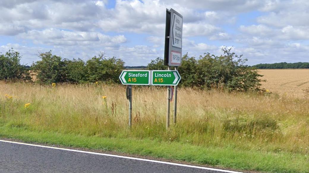 Green road sign at the side of the A15 directing people either towards Sleaford or Lincoln. There is a section of road in view, along with a hedge and a field in the background.