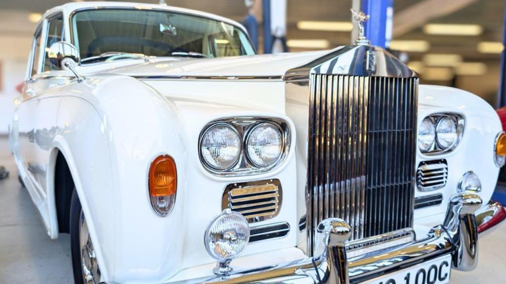 The front of a white Rolls Royce in an indoor space with a large silver grill and the Rolls Royce symbol on top of it.