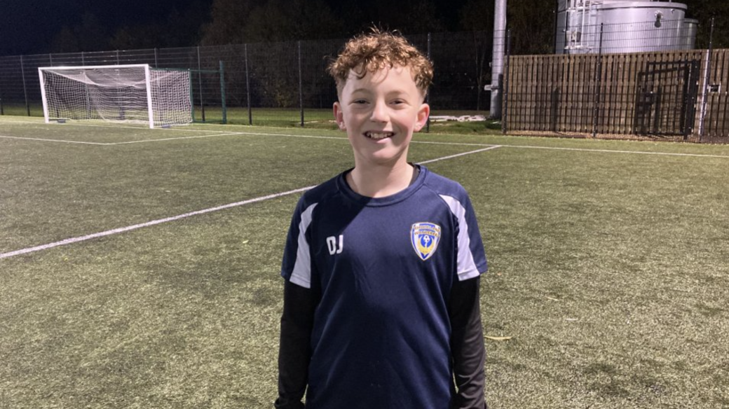 Diesel, 12, smiling at the camera in his navy football club top. He has light brown curly hair and is standing on a football pitch with a goal behind him.