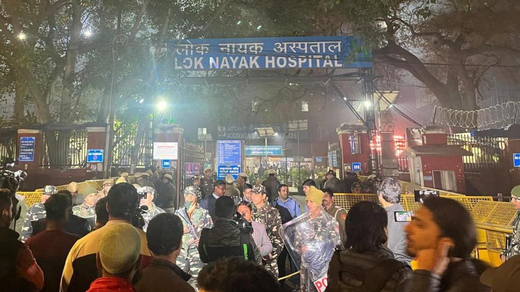 A blurry late night image of the Lok Nayak Hospital, with crowds in the foreground and police personnel guarding the barricade. 