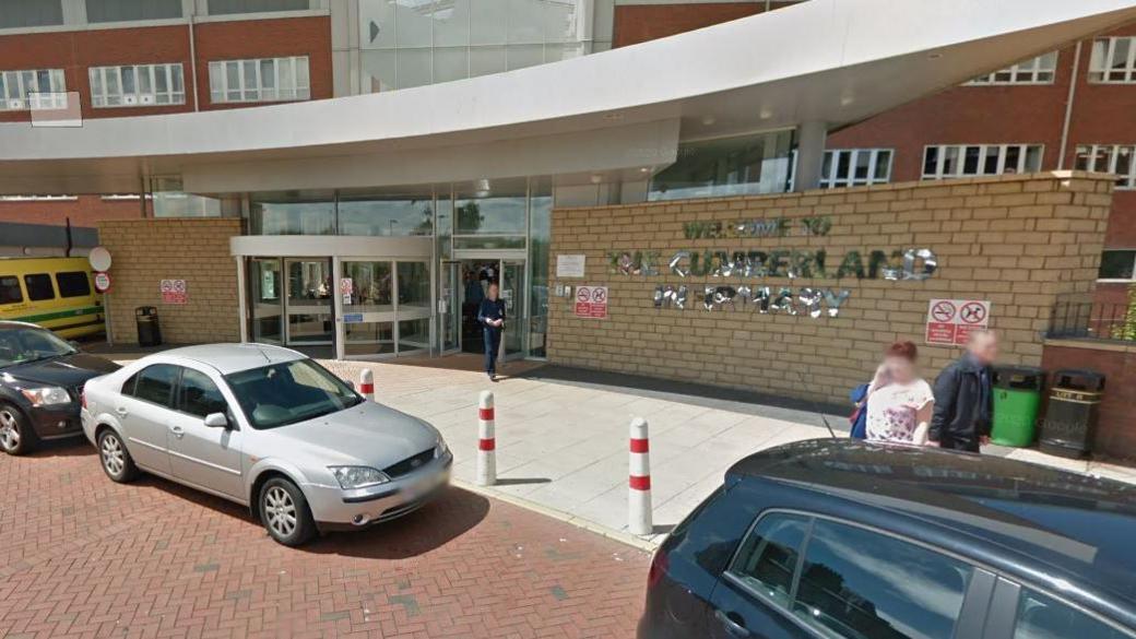 The main entrance at Cumberland Infirmary. A large sign on a brick wall reads "Welcome to the Cumberland Infirmary". Several cars are parked in front and people are entering and leaving the building. There is a curved, silver grey canopy above the entrance, behind which sits the modern red brick hospital building.