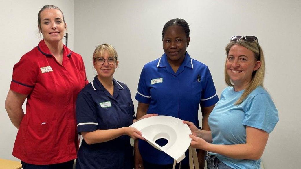 Four women stand together, three wear nurses uniforms. One is red, another is navy and a third is a royal blue. Another woman wears a blue T-shirt and jeans, with sunglasses on her head. Two of the women hold the cradle, which is made of white plastic and has a semi-circular shape.