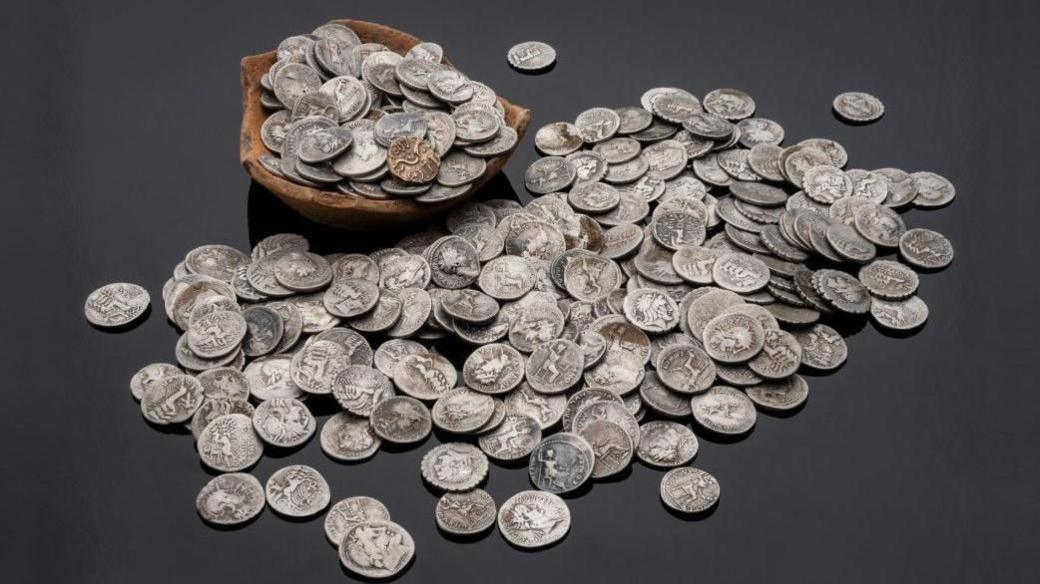 A pile of old-looking silver coins in front of and in a broken pot.