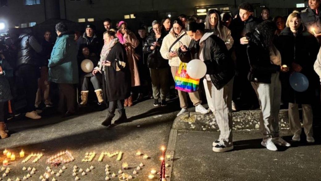 People stand near tributes to Marcus Meade, on the ground, in the dark. Properties are seen in the background of the photo.