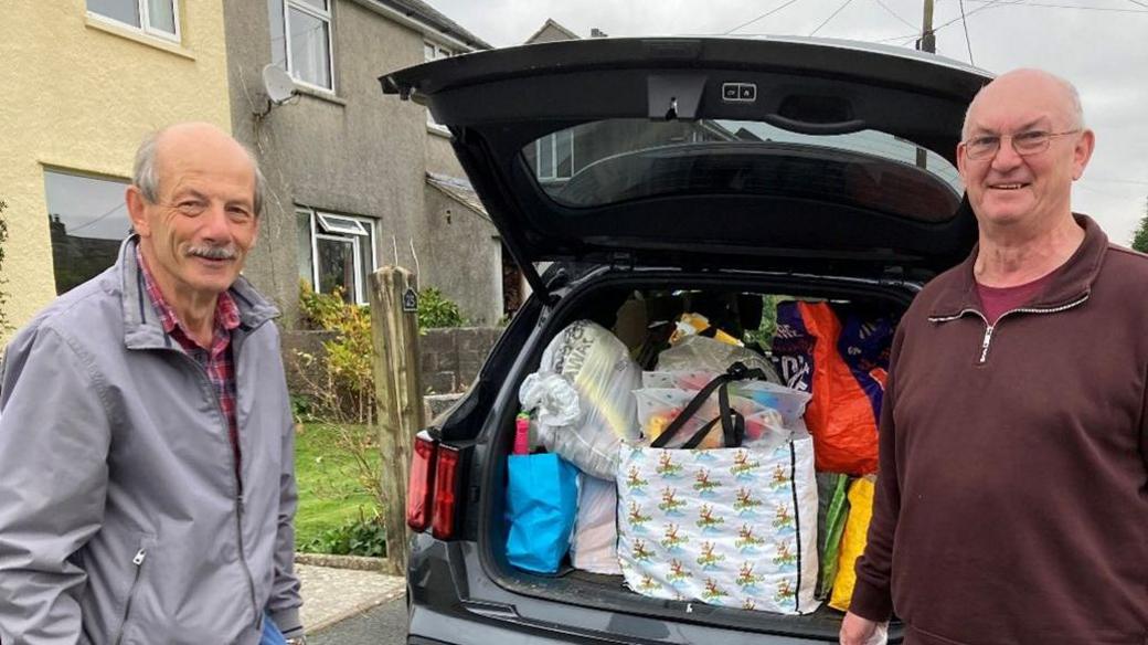 Wally Lake wearing a grey anorak on the left and Nigel Ellis wearing a burgundy jumper on the right. They are both stood in front of a boot full of donations 