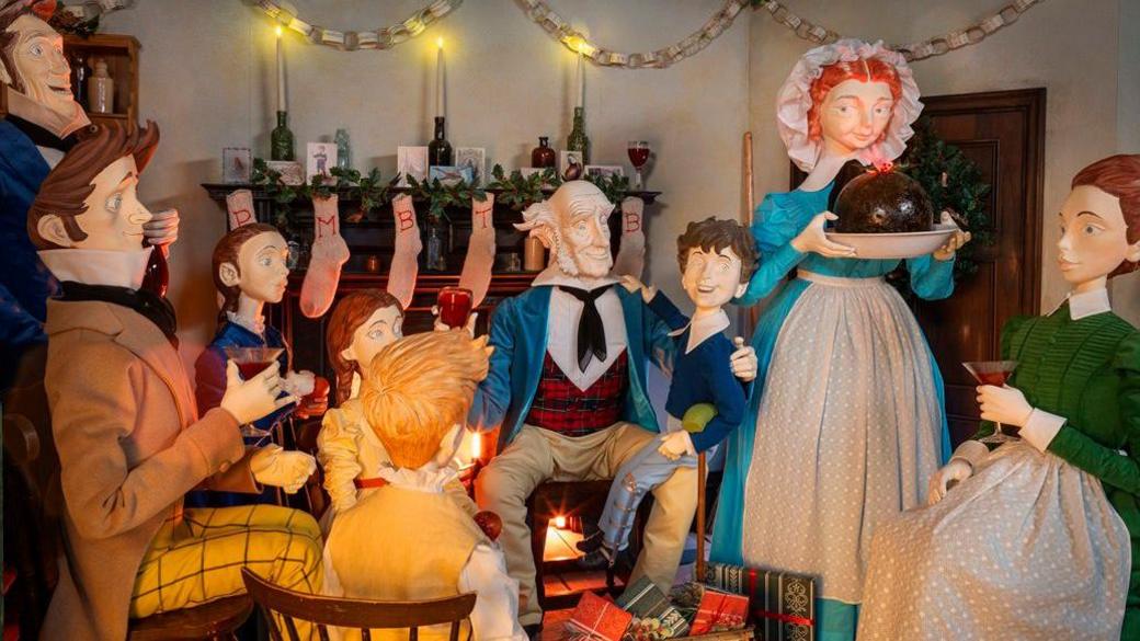 Figurines sat around a fire. A woman holds a Christmas pudding. There are stockings above the fireplace. Scrooge is sat with the family.