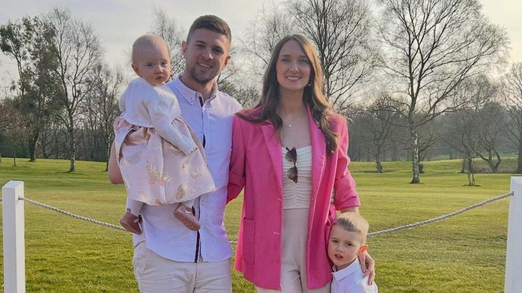 A man and a woman stand smiling in front of a field. The man is holding a young girl wearing a pink dress and the woman has his arm around a young boy who is standing next to her leaning against her leg.