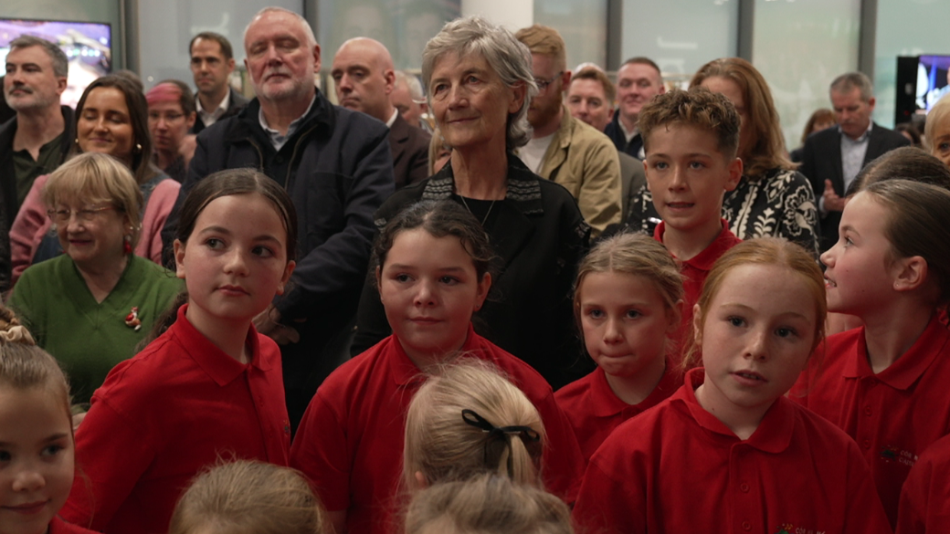 Catherine Connolly stands among a crowd at the event with school children in front of her wearing a red uniform. She has short grey hair and is wearing black clothing. There is other adults surrounding her.