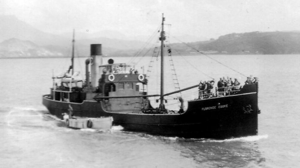 The SS Florence Cooke ship pictured in black and white with mountains in the background. The ship has people stood at the front of the ship. 