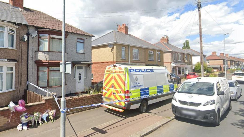 A street view of Scarlet's house, a semi detached two storey house on a street of homes. Half a dozen or so bunches of flowers and balloons have been rested against a garden wall and there are two large police vans parked outside, with a cordon set up with blue and white police tape.