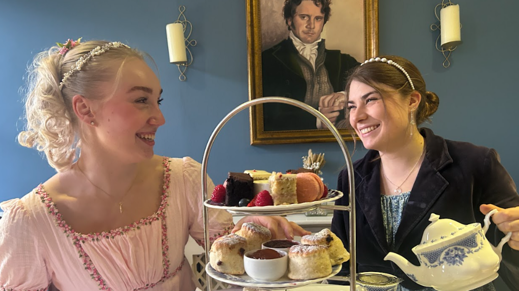 Two ladies dressed in eighteenth century costume smiling at each other in a tea room with a large three-tiered plate of cakes and one of them pouring tea from a china pot. There is a painting of Colin Firth as Mr Darcy on the wall in the background