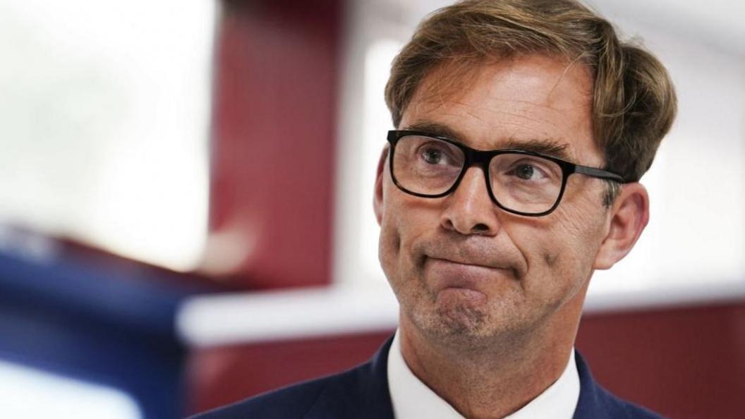 A close-up of Tobias Ellwood, with short brown hair, stubble and glasses, wearing a dark blue suit and white shirt. He has a glum look on his face