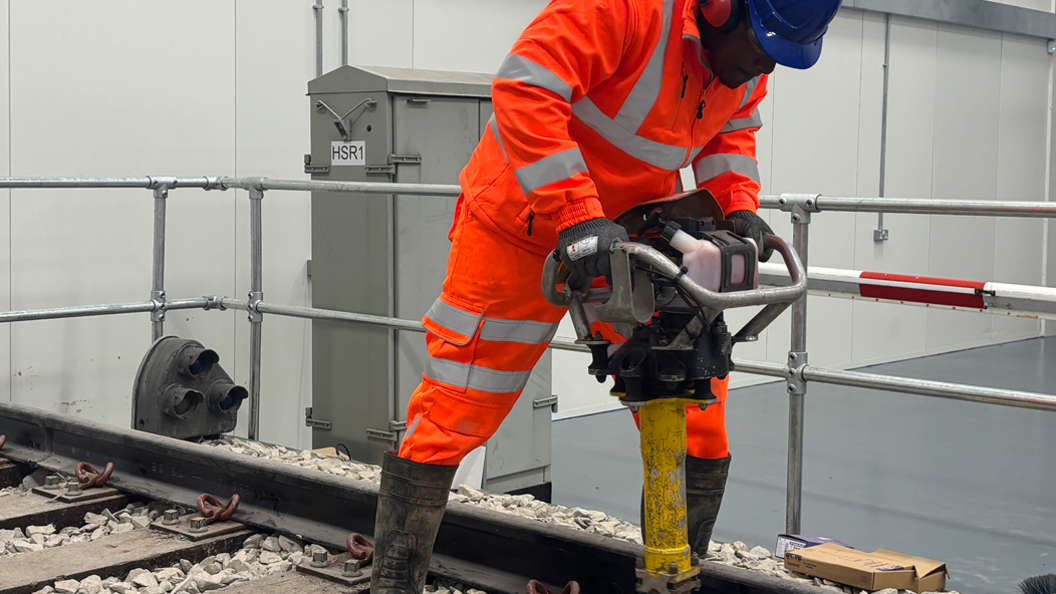 A man in an orange safety suit uses a large drill to work on train tracks. He has a blue hard hat on and wellington boots.