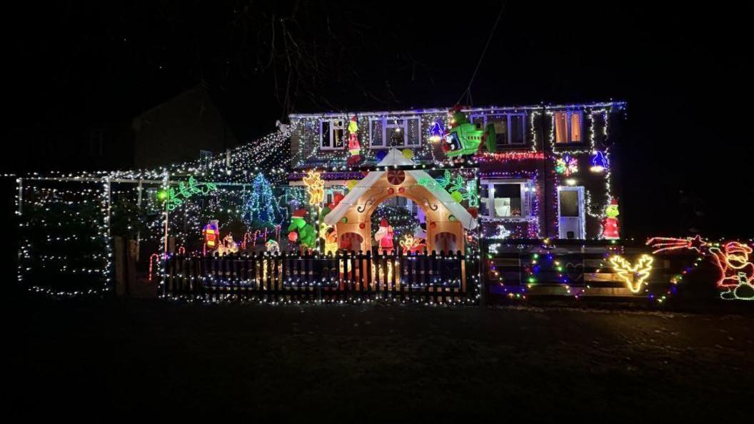 A view from the front of the Christmas display. It is dark and you can see dozens of white and multi-coloured lights surrounding festive displays including a reindeer, a Santa, a Christmas tree and a blow-up archway in the middle.