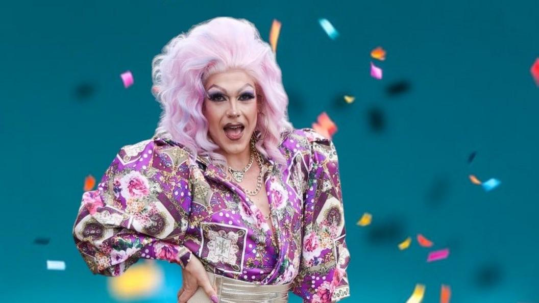 A drag act wearing a bright pink wig, a few necklaces and a purple top. 