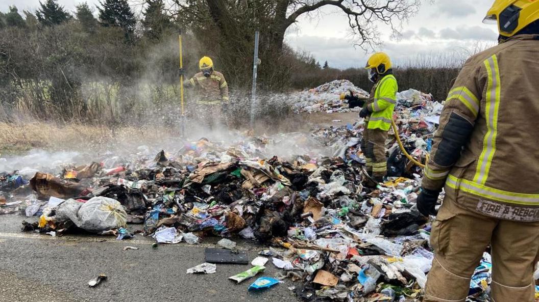 The photo shows three firemen among piles of rubbish. They have a hose, and have put out a fire. A lot of the rubbish has been burnt, and there is smoke above it.