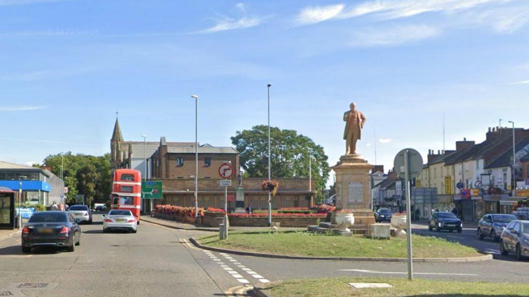 Abington Square, Northampton, showing statue of a man on a plinth (Charles Bradlaugh), which is standing on a grass island. To the left is a blue canopy of a car showroom or similar, there is a church in the centre of the picture and shops with flats above to the right. There are some cars and a red double decker bus on the road to the left.