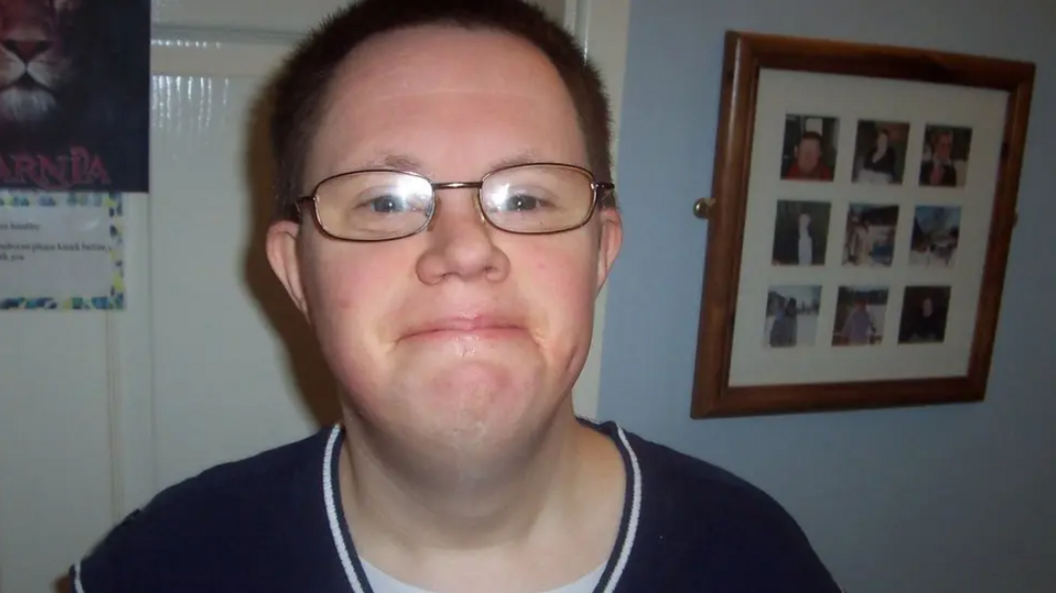 A close-up image of Richard Handley, who is looking directly into the camera. He is smiling and wearing glasses. He is wearing a navy top and is photographed inside a room with a photo collage behind him hanging on a wall. 