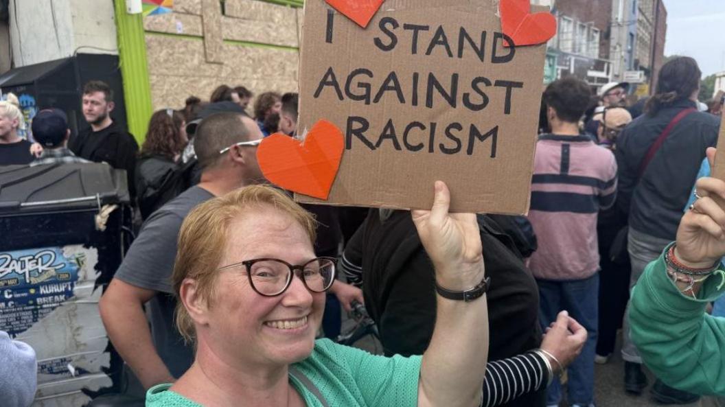 A lady in a green top at a rally in Bristol holding a sign that says 'I stand against racism'. There are also three red hearts on the sign. 