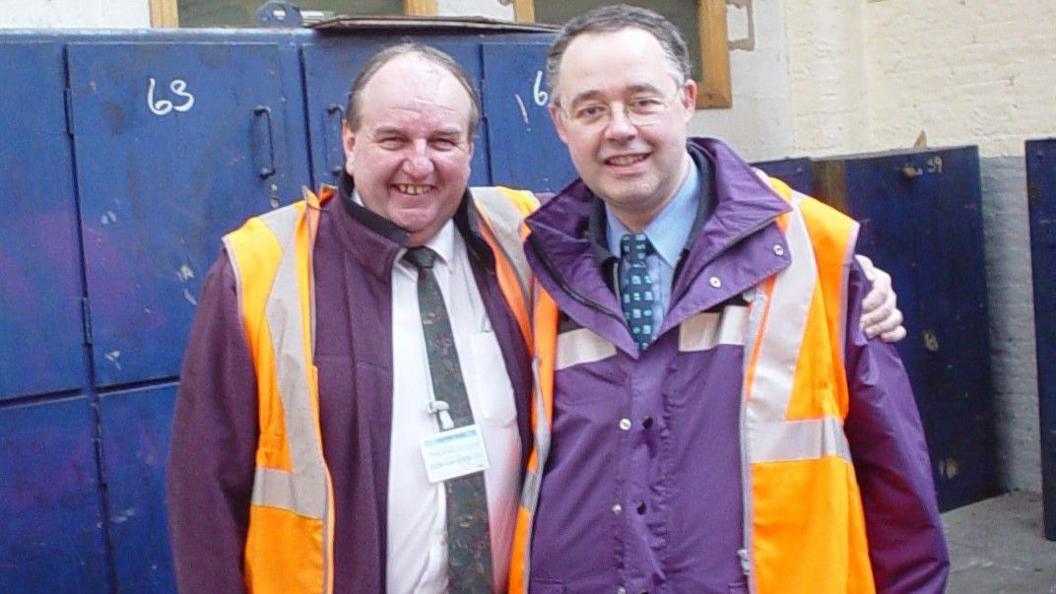 Mr Coffey wearing a purple coat and orange high visibility jacket with another man in similar dress