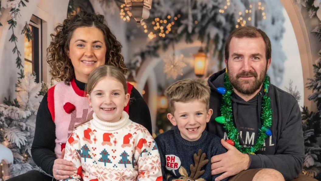 A family photo of Lizzie, Libby, Lewis and Alex who are all wearing Christmas jumpers. They are sat in front of a winter backdrop and are all smiling at the camera. 