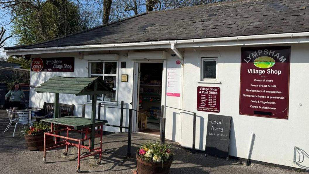 A view of the front entrance to the village shop. The shop has a sign on the front branded with  'Post Office'. The small building is painted white.