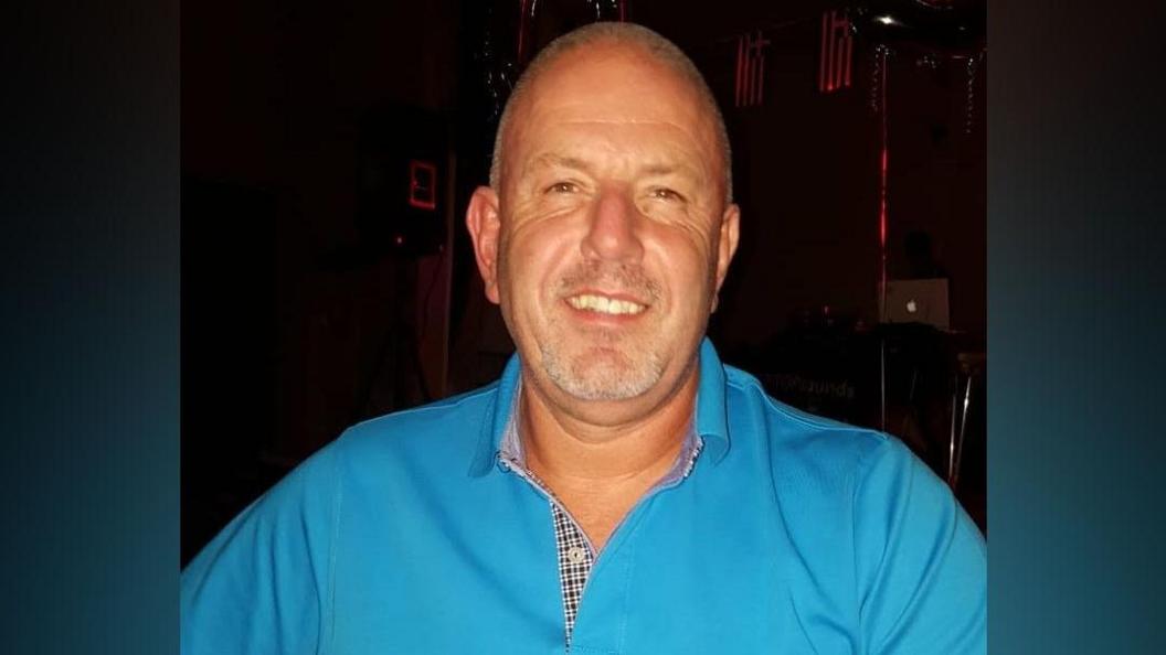 A tanned man in a blue shirt smiling at the camera. He has close cropped graying hair. He is in front of a dark background.