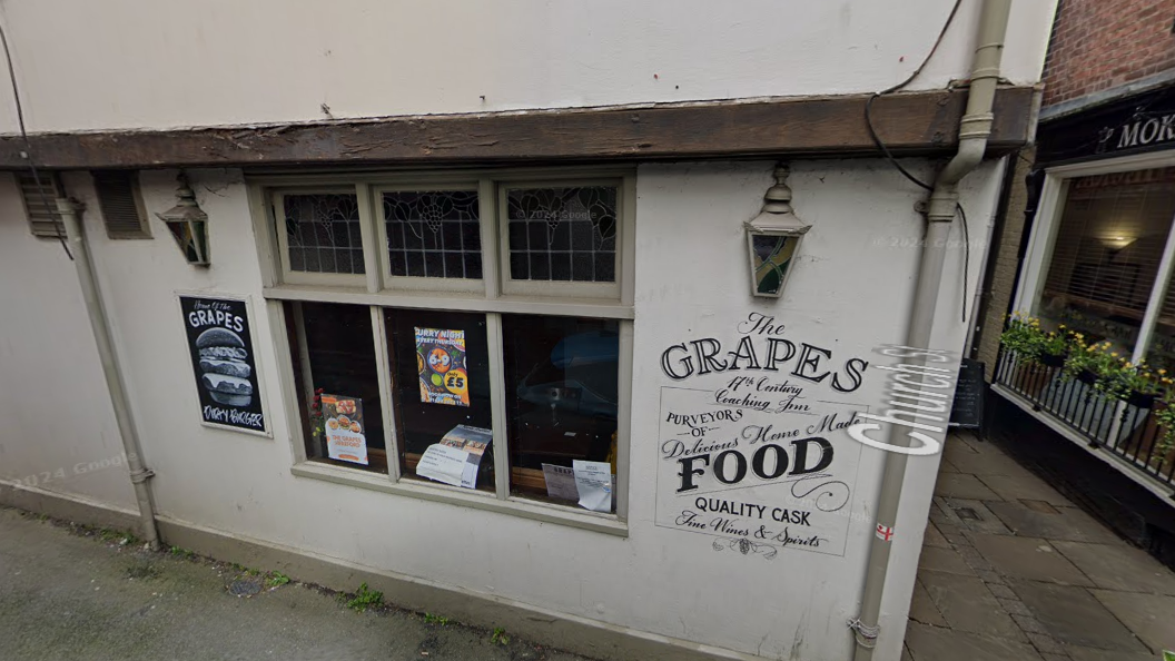 The outside of the Grapes Tavern. It is a white building with the name printed on the side in black lettering. The window contains posters for events, and there is an advert for burgers to the left of the window.