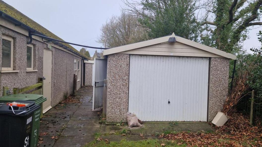 A garage in pale pebble-dash with a white door is on the right of the image. To the left is the side wall, also pebble-dashed, of a church hall.