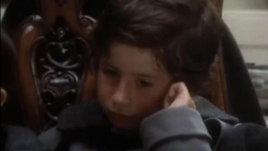 A close up shot of John Oliver as a child, dressed in a grey oversized coat and against a wooden ornamental chair