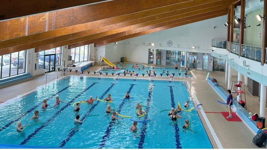 A group of swimmers wearing hats and using flotation aids participate in an aqua fit class, with an instructor standing on the poolside. There is a second, smaller pool beyond it.