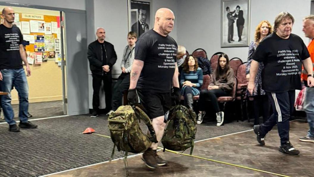 A bald man in a black T-shirt and black shorts is carrying two green camo print backpacks, and he is walking with them. There are people spectating around him