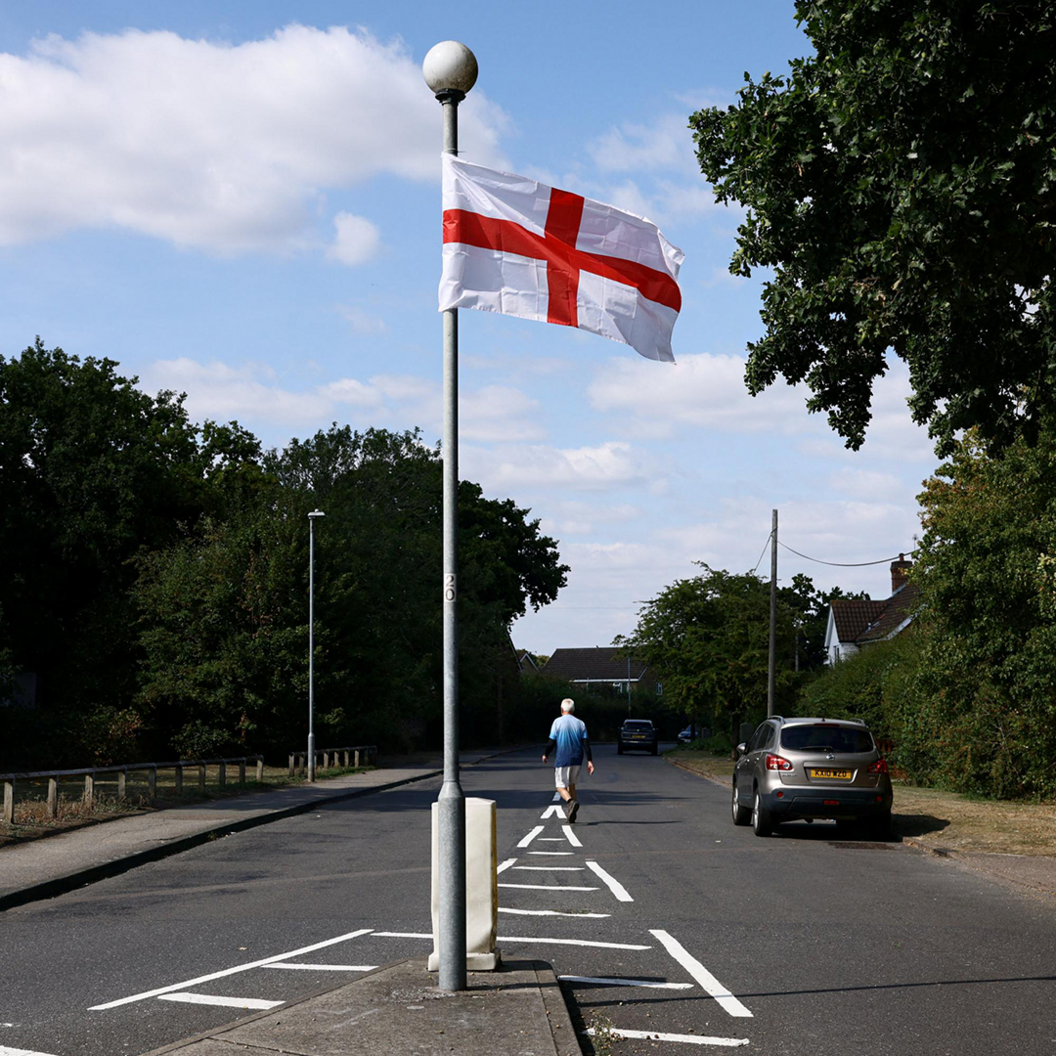 A flag of St George hangs from a lamppost on an island in the middle of an empty road, as a man walks down the road in the background.