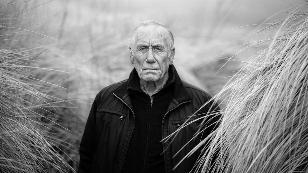 A black and white photo of Richard Lee. He is wearing a black jacket and jumper and is surrounded by tall weeds.