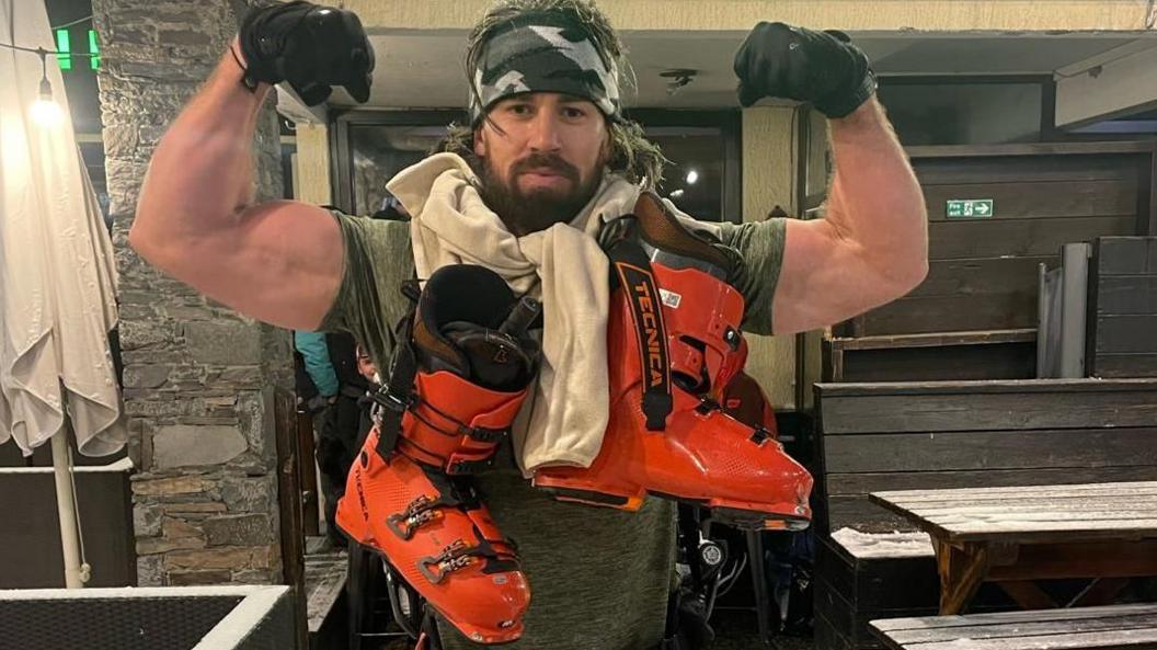 Mike Humphreys wearing a grey and white bandana around his forehead is flexing his muscles for the camera. He is wearing black gloves and has a pair of bright orange ski boots around his neck.