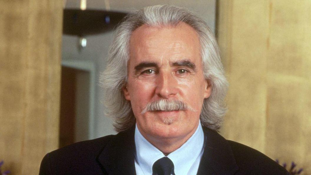Charlie Miller has shoulder-length silvery hair and moustache. He is wearing a black suit jacket, white shirt and tie.