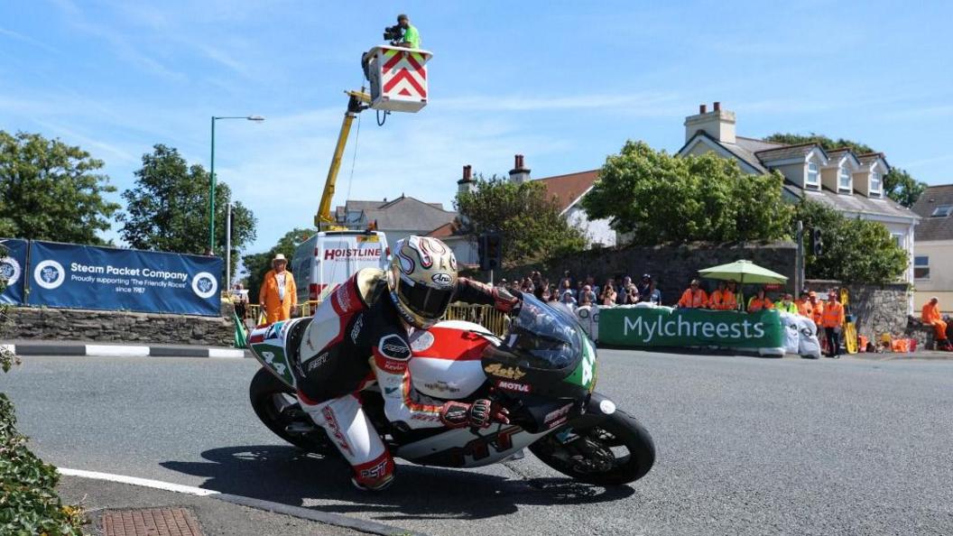 Rob Hodson leaning to the left with a cameraman in the air in a cherry picker in the background.